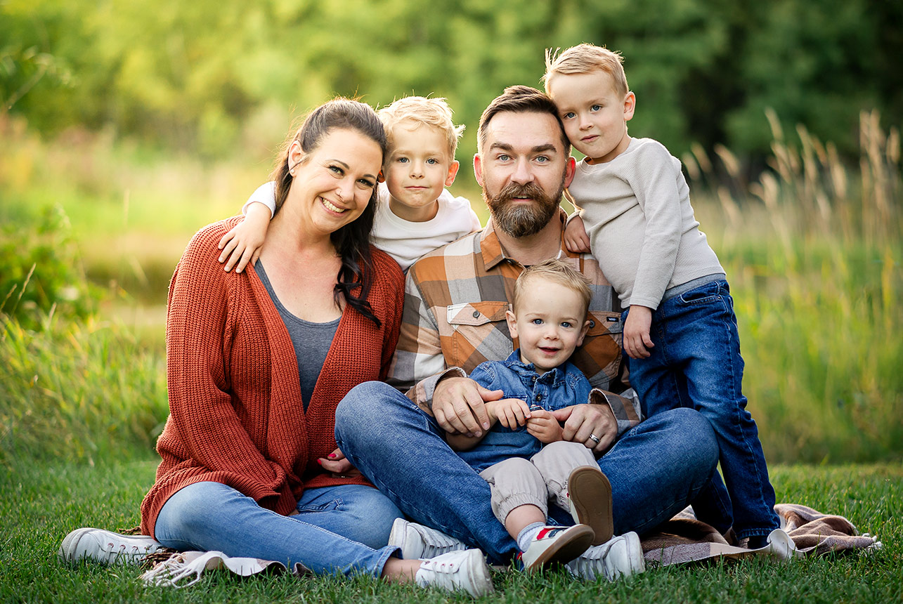 Family of two parents and three boys on grass at golden hour, toddler in father’s lap and older brothers leaning on parents