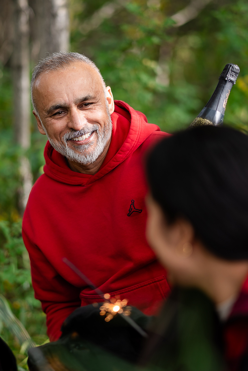 Smiling man in a red hoodie enjoys a forest picnic with friends, champagne bottle and sparklers highlighting the celebration near Edmonton