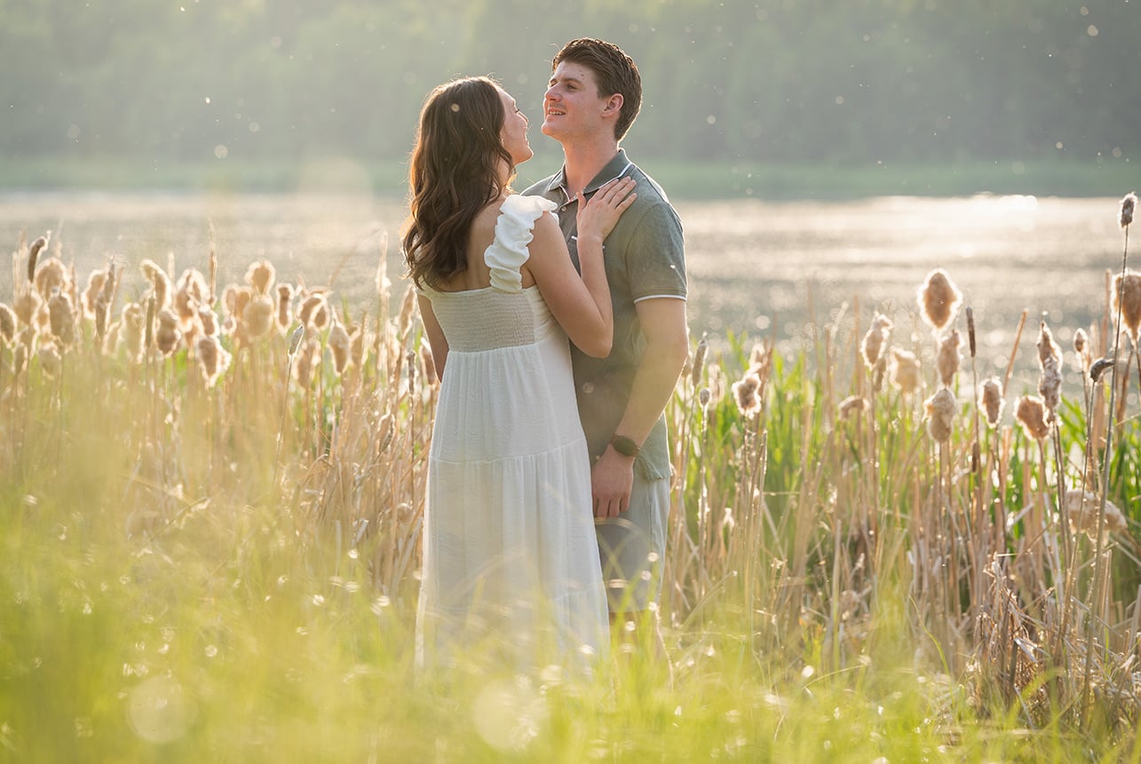 Couple in summer attire standing close in a field of cattails beside a shimmering lake