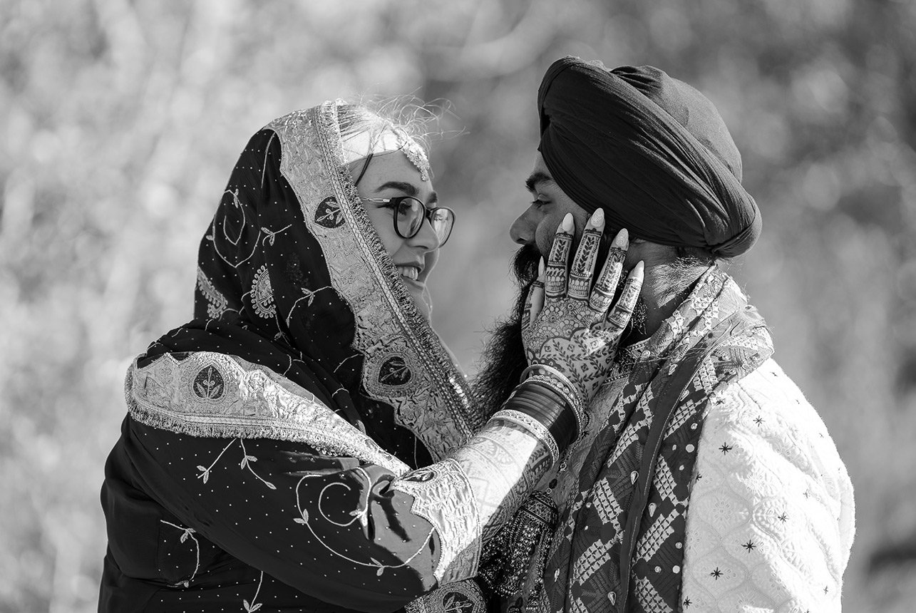 Bride in traditional Sikh attire holds groom’s face, both smiling lovingly