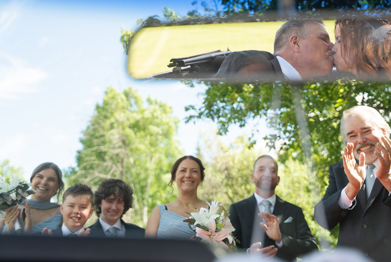 Wedding party cheers as bride and groom kiss in car rearview mirror