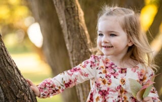 Smiling young girl in a floral dress standing by a tree, holding autumn leaves during a golden hour photo session in Sherwood Park, captured by Catriona Hope Photography