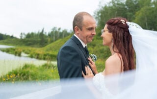 Bride and groom face to face by the water, veil drifting through the foreground