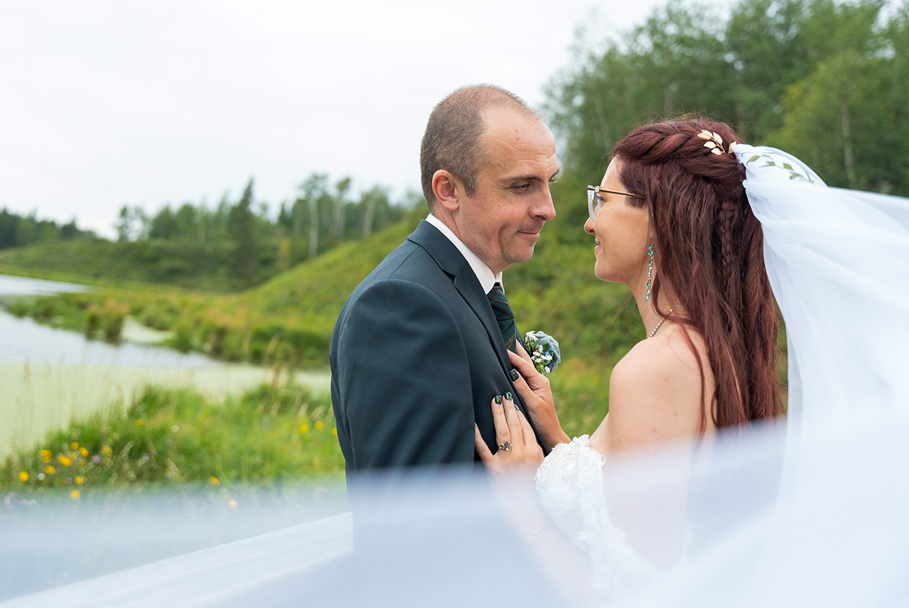 Bride and groom face to face by the water, veil drifting through the foreground