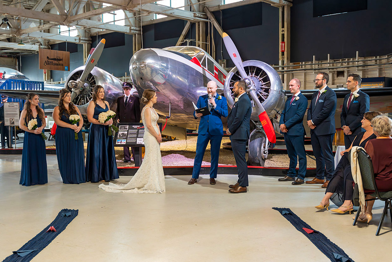 Bride and groom exchange vows at the Alberta Aviation Museum with a polished vintage aircraft behind them, wedding party lined up on both sides