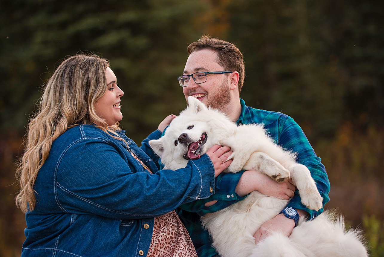 Couple smiling and cuddling with their fluffy white dog during an outdoor engagement session