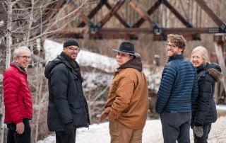 Family walking together on a snowy trail and looking back near a rustic bridge during a natural outdoor winter photo session
