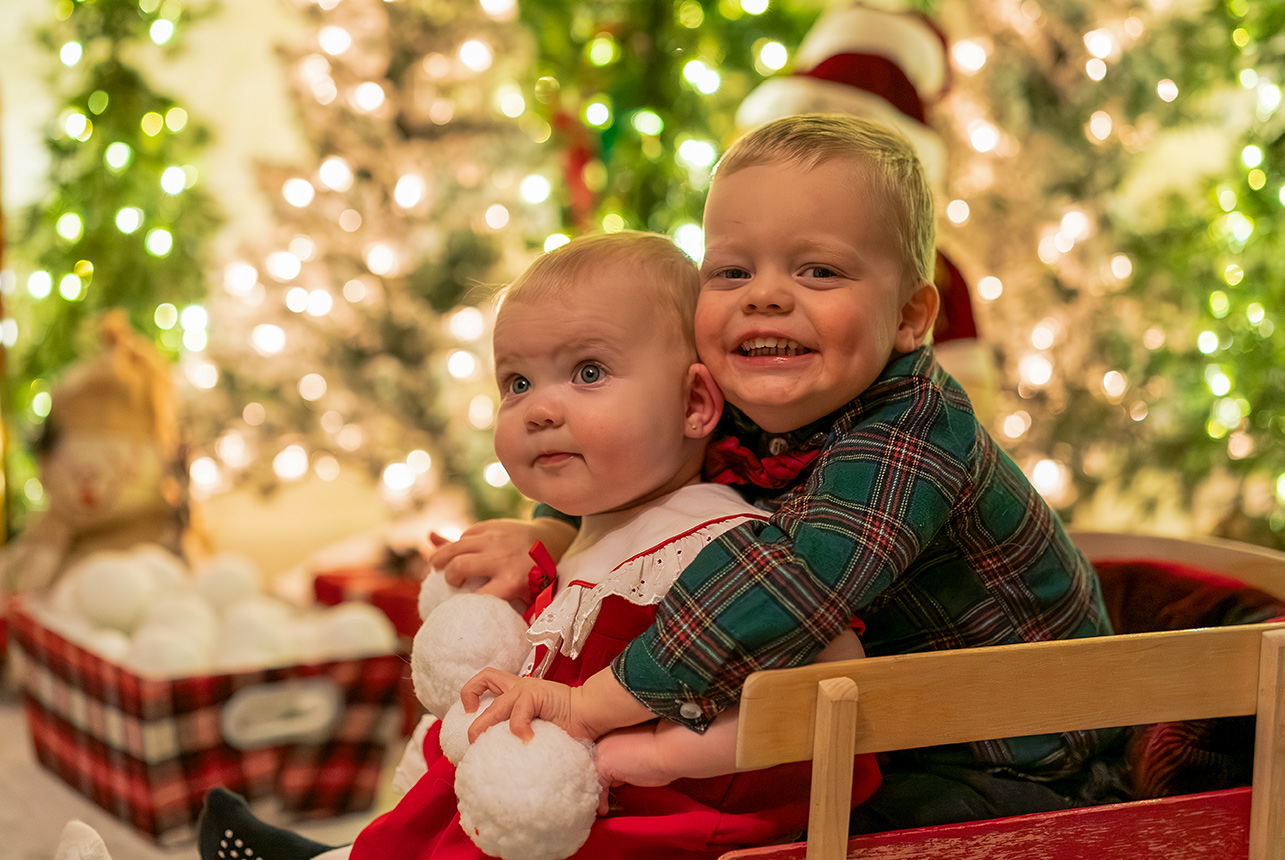 Smiling boy hugging a baby holding a white teddy bear in front of glowing Christmas trees