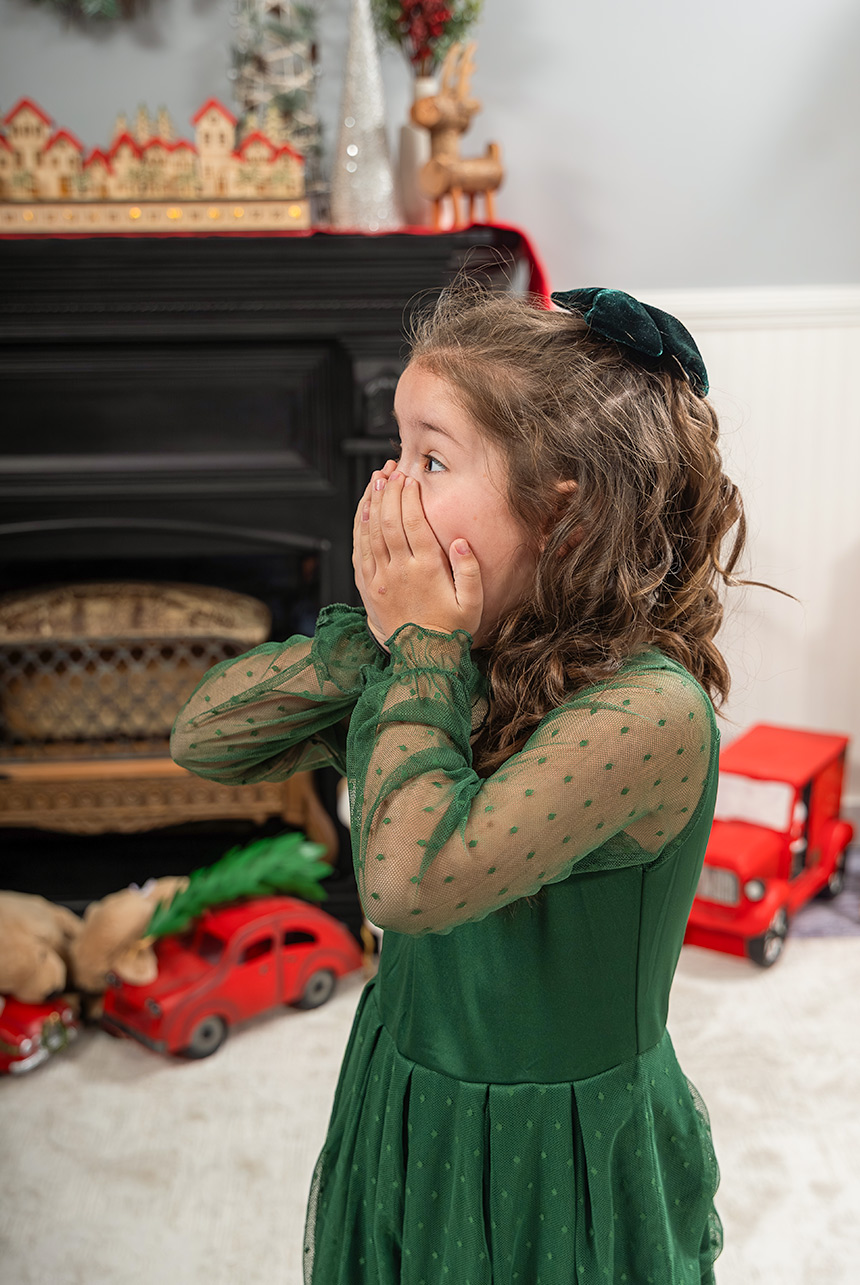 Girl in a green dress covering her mouth beside a decorated Christmas fireplace and miniature red vehicles