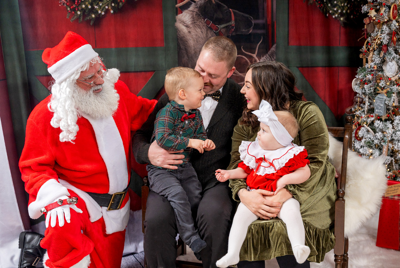 Family of four sitting with Santa in front of a red barn backdrop and a decorated Christmas tree